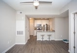 This single-floor space showcases a thoughtfully designed open-concept layout that connects the kitchen and living areas seamlessly. The kitchen features a peninsula-style breakfast bar with granite countertops and seating for two bar stools, making it an ideal spot for casual dining or entertaining. A sink with modern fixtures is visible along the counter, complemented by cabinetry and what appears to be standard appliances. The main living area is finished with rich, dark hardwood-style flooring that adds warmth and visual depth to the space. Neutral gray walls create a clean, versatile backdrop throughout. A doorway with a classic paneled white door leads to an adjoining carpeted room, offering a clear distinction between living and private spaces. The ceilings feature a subtle tray detail with a pendant light fixture, adding a touch of architectural interest. White trim and baseboards provide crisp contrast against the neutral wall tones. The overall ambiance is modern and understated, with a practical flow between areas that makes the space feel cohesive and well-organized.