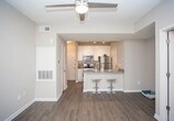 This single-floor space showcases a thoughtfully designed open-concept layout that connects the kitchen and living areas seamlessly. The kitchen features a peninsula-style breakfast bar with granite countertops and seating for two bar stools, making it an ideal spot for casual dining or entertaining. A sink with modern fixtures is visible along the counter, complemented by cabinetry and what appears to be standard appliances. The main living area is finished with rich, dark hardwood-style flooring that adds warmth and visual depth to the space. Neutral gray walls create a clean, versatile backdrop throughout. A doorway with a classic paneled white door leads to an adjoining carpeted room, offering a clear distinction between living and private spaces. The ceilings feature a subtle tray detail with a pendant light fixture, adding a touch of architectural interest. White trim and baseboards provide crisp contrast against the neutral wall tones. The overall ambiance is modern and understated, with a practical flow between areas that makes the space feel cohesive and well-organized.