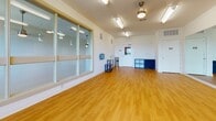 Located at 615 Rio Rd E in Charlottesville, Virginia, this single-floor studio space is thoughtfully designed to support a variety of movement-based activities, from dance and yoga to fitness instruction. The open floor plan is anchored by warm-toned hardwood flooring that extends throughout the space, providing a clean, resilient surface well-suited for active use. A wall of large, floor-to-ceiling mirrors lines one side of the studio, enhancing the sense of depth and allowing participants and instructors to monitor form and alignment with ease. A mounted flat-screen display is positioned alongside the mirror wall, supporting instructional or media use. Natural light fills the room through multiple windows that frame views of the surrounding Charlottesville landscape, creating an inviting and energizing atmosphere. Overhead fluorescent lighting and a ceiling fan with a light fixture ensure the space remains well-lit and comfortable regardless of the time of day. Interior glass panel windows connect the studio to an adjacent area, maintaining visual openness while offering a degree of separation. The space also includes a keyed entry door with a window, built-in storage cubbies, and utility fixtures such as a sanitizer dispenser and waste receptacle, reflecting practical, user-focused design throughout.
