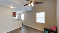 This single-floor residence offers a well-designed, open-concept layout that maximizes both functionality and comfort. The main living area is anchored by rich, dark hardwood-style flooring that extends throughout, providing a warm and cohesive aesthetic. Vaulted ceilings elevate the sense of space, while a ceiling fan with a built-in light fixture ensures year-round comfort and ambient illumination. Two windows fitted with white horizontal blinds allow natural light to filter in, brightening the neutral, beige-toned walls that create a clean and welcoming atmosphere. A textured ceiling adds a subtle design detail overhead. The living area flows naturally into a kitchen pass-through, where rich cherry-toned cabinetry and a granite-style countertop are visible, offering a stylish and practical culinary space. A wall-mounted HVAC unit provides efficient climate control directly within the living area. The overall ambiance is light, airy, and move-in ready, with a straightforward layout that lends itself well to a variety of furniture arrangements and personal styles. This space combines practical features with warm finishes to create an inviting, comfortable home environment.