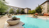 The pool and grill areas form a thoughtfully designed outdoor amenity complex set within a courtyard framed by multi-story residential buildings. The centerpiece is a generously sized resort-style swimming pool with clear turquoise water, a wading shelf entry, and tiled accent details. The pool deck is finished in light-toned concrete and furnished with rows of lounge chairs, while large decorative concrete planters filled with agave and ornamental grasses add structured greenery throughout.

Adjacent to the pool, a dedicated grill zone features multiple stainless steel grills positioned along a landscaped pathway lined with native plantings and ornamental grasses. Overhead string lights lend an inviting ambiance to the area during evening hours. Nearby, an outdoor lounge and dining patio offers weather-resistant seating arrangements, including sectional sofas with vibrant cushions and dining tables with chairs, ideal for social gatherings.

Connecting pathways wind through manicured lawn areas shaded by mature trees, creating a park-like atmosphere. Additional sun loungers are positioned along secondary walkways bordered by layered landscaping. Accessible entry points with ramps integrate seamlessly into the overall design, contributing to a cohesive, well-appointed outdoor living environment.