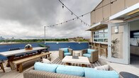 Located at 616 Oberlin Rd in Raleigh, NC, this rooftop amenity space offers an exceptional blend of indoor and outdoor living designed for gathering and entertaining. The expansive open-air terrace is the centerpiece of the space, featuring large-format tile pavers, wicker lounge seating with aqua-toned cushions, a coffee table, and a dining table with bench seating — all set beneath charming overhead string lights that create an inviting ambiance day or night. Bold blue paneled perimeter walls frame sweeping panoramic views of the Raleigh skyline and surrounding tree canopy, making this terrace a standout feature. A covered interior area connects seamlessly to the rooftop via a roll-up garage-style door, offering flexible indoor-outdoor flow. Inside, a sleek kitchenette is equipped with light wood cabinetry, dark granite countertops, a stainless steel sink, a refrigerator, and a wall-mounted television — ideal for hosting events. The interior also includes a fully tiled restroom with grab bars and a neutral tile surround. Light wood-look flooring throughout the interior ties the space together with a clean, contemporary aesthetic. This well-appointed rooftop venue is thoughtfully designed for both casual gatherings and more formal events.