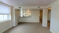 This single-floor space features an open-concept design that seamlessly connects the living area to the kitchen, creating a bright and functional layout. The living area is finished with neutral-toned carpet, lending a comfortable and understated feel to the space. A structural half-wall with a dark-topped peninsula separates the kitchen from the living area while maintaining an open sightline throughout.

The kitchen is well-appointed with crisp white cabinetry that extends to the upper walls, providing ample storage. The dark countertop of the peninsula offers a contrasting design element, adding visual interest to the otherwise light interior. A window in the kitchen allows for natural light to enter the cooking area.

The walls throughout are painted in a clean, bright white, contributing to an airy and open atmosphere. Ceiling-mounted HVAC vents are integrated into the ceiling, ensuring climate control throughout the space. A hallway visible from the main living area suggests additional rooms beyond, further extending the functional footprint of this well-organized, single-level layout.