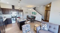 This well-appointed two-story home offers a thoughtfully designed open-concept main floor that seamlessly integrates the kitchen, dining, and living areas into a cohesive and inviting space. The kitchen is a standout feature, showcasing rich dark cabinetry, stainless steel appliances including a refrigerator and range, and a granite-topped island with bar seating — perfect for casual dining or entertaining. Pendant lighting above the island adds a modern touch, while a chandelier over the dining area creates a warm, welcoming ambiance. The dining space comfortably accommodates a full table set with bench seating, ideal for gatherings. The living area flows naturally from the dining space, offering a relaxed setting with accent chairs and ample room to unwind. Light wood-toned flooring runs throughout the main level, enhancing the bright and airy feel of the space. A staircase leads to the upper floor, adding to the home's multi-level functionality. The overall design balances contemporary style with practical living, making it a comfortable and visually appealing residence suited for everyday life.