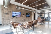 Located in Waukee, Iowa, this single-floor amenity center offers a generously sized, thoughtfully designed space that blends modern industrial aesthetics with comfortable, functional living areas. The open lounge area serves as the heart of the facility, featuring exposed brick accent walls, warm wood-paneled ceiling insets with integrated linear lighting, and polished concrete floors. Leather seating arrangements and area rugs create inviting gathering zones, while a snack and beverage bar with under-counter refrigeration adds everyday convenience. Dual flat-screen TVs mounted on the brick wall anchor a casual entertainment area nearby. A glass-enclosed conference or workspace room provides a quieter setting for focused activities. The fitness center is divided into distinct zones: one area houses treadmills, ellipticals, and weight equipment alongside exposed masonry walls and multiple mounted screens, while a second open exercise room with rubberized flooring, a ceiling fan, and abundant natural light accommodates a variety of workout routines. A dining area with high-top tables and bar seating offers a casual space for meals or socializing, complemented by a kitchenette with full-size appliances. Natural light flows throughout via large windows, and exposed ductwork contributes to the space's contemporary industrial character.