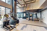 This upscale fitness facility offers a thoughtfully designed, generously sized workout environment spread across a single floor. The main gym area commands attention with soaring double-height ceilings, exposed wood-beam architectural detailing, and dramatic pendant and hoop lighting that lend the space a contemporary yet warm character. Large floor-to-ceiling windows along one wall flood the area with natural light while providing open views of the surrounding courtyard, creating an energizing atmosphere. The flooring features a stylish combination of light wood-look planks and geometric patterned tile insets, seamlessly transitioning between zones.

The main gym is comprehensively equipped with cardio machines including treadmills, ellipticals, and a stair climber, alongside a full cable machine, a multi-functional strength rack, a dumbbell rack, kettlebells, medicine balls, and a functional training rig. Floor-to-ceiling mirrors amplify the sense of space. A water station and cabinetry are conveniently tucked near the entry.

A dedicated studio room provides a quieter, focused space for yoga, stretching, or group classes, featuring warm hardwood-style flooring, full-wall mirrors with a ballet barre, built-in storage for yoga mats and props, a wall-mounted display screen, and recessed lighting. Together, these two areas form a complete and refined fitness destination.