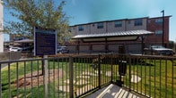 This bark park is a thoughtfully designed outdoor amenity situated within a multi-story residential community. The space is fully enclosed by sturdy dark metal ornamental fencing, providing a secure environment for pets to roam freely. A gated entry with a self-latching mechanism ensures safe access, while a prominent "Bark Park" rules and information signage panel is posted at the entrance for resident reference.

The ground surface consists of maintained turfgrass interspersed with stepping stone pathways, offering a clean and functional play area. A low natural stone retaining wall borders one side of the park, adding a structured landscape element that separates the dog park from the adjacent covered parking area. Overhead shade structures with metal panel roofing are positioned nearby, providing shelter from direct sunlight. Young trees are planted within and around the perimeter, contributing shade and greenery over time.

The surrounding multi-story residential buildings with brick and stucco exteriors frame the space, giving the park a well-integrated community feel. The overall ambiance is open, bright, and practical, designed to accommodate pets and their owners in a safe, accessible outdoor setting.