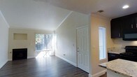 This single-floor residence offers a welcoming and well-lit living environment defined by clean lines and thoughtful architectural details. The living area immediately impresses with its vaulted ceiling, which lends a sense of openness and volume to the space. Crown molding runs throughout, adding a refined finishing touch to the warm, neutral-toned walls.

Rich, dark hardwood-style flooring extends across the main living area, providing a stylish and durable foundation that contrasts beautifully with the bright, white trim and baseboards. Large windows dressed with vertical blinds invite an abundance of natural light, creating an airy and cheerful atmosphere while offering views of the surrounding greenery outdoors.

A standout feature of the living room is the classic fireplace, framed in sleek black, which serves as a natural focal point. Above it, a recessed niche in the wall provides a charming display area. The front entry is flanked by a sidelight window, enhancing the sense of openness upon arrival. A textured ceiling in the entryway transitions to the smooth vaulted ceiling of the main living space, adding architectural interest. Overall, the space presents a comfortable and inviting environment with character-rich details throughout.