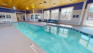 Located at 200 Edinborough Dr in Durham, NC, this single-story indoor aquatic facility offers a well-appointed swimming environment designed for both recreation and relaxation. The centerpiece is a generous lap pool with clearly marked lanes, complemented by a separate spa with stepped entry, both finished in vibrant aqua tones. The space is crowned by a warm, natural wood-planked ceiling that lends a distinctive architectural character, contrasting elegantly with the bold navy blue upper walls and crisp white wainscoting below. Abundant natural light pours in through large, multi-pane windows positioned around the perimeter, offering views of the surrounding landscaped grounds and creating an open, airy atmosphere throughout. Scattered around the pool deck are modern metal-framed seating arrangements upholstered in blue-striped and solid blue cushions, providing comfortable lounging areas for guests. A dedicated observation and lounge area with floor-to-ceiling windows overlooks both the indoor pool and an outdoor pool area, making it a versatile social space. Safety signage, a wall-mounted life ring, and poolside equipment are thoughtfully integrated throughout. The smooth concrete pool deck and well-maintained finishes reflect a commitment to both functionality and comfort in this inviting aquatic retreat.