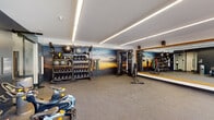 This single-floor fitness center is thoughtfully designed to accommodate a full range of workout disciplines within a spacious, open layout. The space is divided into distinct functional zones, each outfitted with professional-grade equipment. The cardio area features multiple Matrix treadmills and elliptical machines positioned along a generous floor-to-ceiling window wall, offering views of landscaped outdoor surroundings and allowing ample natural light to fill the room. A row of Matrix cycling bikes occupies a dedicated section, complemented by a glass partition that subtly separates training areas while maintaining an open feel throughout.

The strength and functional training zone is anchored by a large, dramatic sunset mural that spans the accent wall, creating a visually dynamic backdrop. A multi-tiered rack holds an extensive selection of kettlebells, medicine balls, and dumbbells, while resistance bands and TRX-style suspension trainers are conveniently accessible nearby. A cable machine system provides versatility for strength training. A floor-to-ceiling mirror with a warm wood-framed border lines one wall, enhancing the sense of openness. Linear recessed LED lighting runs across the white ceiling throughout, providing bright, even illumination. Durable speckled carpet and rubber flooring complete the functional, modern aesthetic of this well-appointed fitness facility.