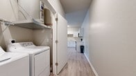 This 870-square-foot, single-floor apartment at 1300 N Lancaster Rd in South Elgin, IL offers a functional and well-appointed two-bedroom, two-bathroom layout. The open living area features light wood-look vinyl plank flooring and a ceiling fan, creating a bright and airy atmosphere with natural light filtering through large windows fitted with horizontal blinds. The kitchen is efficiently designed in an L-shape with white cabinetry, white countertops, and a full suite of black appliances including a refrigerator, electric range, microwave, and dishwasher, all set atop matching wood-look flooring. Both bedrooms are carpeted and generously lit by large double windows. The primary bedroom measures 130 square feet, includes a ceiling fan, and connects directly to a full bathroom featuring a tub/shower combo, white vanity with cabinet storage, and wood-look flooring. A second bathroom offers a similar vanity setup with lighting above a large mirror. A dedicated walk-in closet with wire shelving provides ample storage. The in-unit laundry area houses a full-size top-loading washer and dryer with additional shelving above, adding everyday convenience to this practical and move-in-ready home.