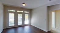 This well-appointed single-floor space offers a clean, modern aesthetic defined by thoughtful design details and quality finishes throughout. The main living area immediately draws attention with its rich, dark hardwood flooring that provides a warm and elegant foundation for the room. Neutral, warm-toned walls create a versatile backdrop that complements a variety of interior styles.

A standout feature of the space is the set of classic French doors flanked by full-length sidelights and transom windows above, flooding the interior with generous natural light while offering a direct connection to the outdoors. White blinds on the doors and windows allow for adjustable privacy and light control. The clean white trim and baseboards throughout provide a crisp contrast to the warm wall tones and dark flooring.

The ceiling is finished with a flush-mount light fixture, and a visible HVAC vent ensures year-round comfort. The space also includes multiple electrical outlets and a wall switch, offering practical utility alongside its aesthetic appeal. Overall, the space presents a bright, open, and move-in-ready environment that balances functional design with inviting visual warmth.
