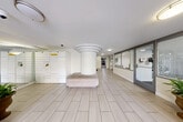This single-floor building lobby presents a clean, functional design finished in light neutral tones throughout. The spacious main lobby area is anchored by a distinctive cylindrical structural column with tiered crown detailing, serving as a visual centerpiece. Light wood-look tile flooring extends across the entirety of the space, contributing to the bright, airy atmosphere enhanced by recessed and flush-mount ceiling lighting.

A dedicated package management station lines one wall, featuring a modern smart locker system alongside a traditional mailbox array, providing convenient mail and parcel retrieval for residents. Adjacent to the lockers, a low upholstered bench offers a comfortable waiting area. A residents-only access door with key-fob entry leads to additional building amenities, framed by floor-to-ceiling glass partitions fitted with horizontal blinds for privacy.

The lobby connects to an elevator bay finished with stone-accented surrounds and stainless steel doors, accompanied by a glass-paned stairwell entry. The covered vehicular entry drive features tiled pavers, textured stucco walls, and integrated planters with lush greenery, offering a welcoming arrival experience with views of the surrounding neighborhood streetscape.
