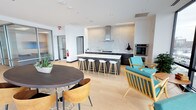 This community lounge is a thoughtfully designed, open-concept social space that seamlessly blends kitchen, dining, and lounge areas into one inviting environment. The space is bathed in natural and recessed lighting, which highlights the light hardwood flooring that runs throughout and contributes to a warm, contemporary ambiance.

At the heart of the lounge is a sleek kitchen island with a striking dark wood finish and a crisp white quartz countertop, lined with modern white bar stools that invite casual seating and conversation. The kitchen is fully equipped with professional-grade stainless steel appliances, including a wall oven, a gas cooktop, a stainless range hood, and a full-size refrigerator — making it ideal for hosting events or community gatherings.

The surrounding walls feature a subtle textured tile backsplash that adds depth and a refined aesthetic to the kitchen zone. Adjacent to the island, a stylish round dining table with mixed-material chairs offers a more intimate setting for smaller groups.

The lounge area incorporates mid-century modern inspired seating, including a distinctive teal accent chair with a wooden frame, adding a bold pop of color against the neutral palette. High ceilings and white walls enhance the sense of openness, while large glass doors provide connectivity to adjacent spaces, completing this well-appointed and versatile community hub.