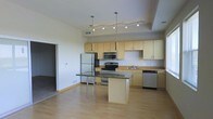 This single-floor space showcases a thoughtfully designed open-concept layout that seamlessly blends the kitchen and living areas. The kitchen is a standout feature, equipped with a full suite of stainless steel appliances, including a refrigerator, electric range, microwave, and dishwasher. Light maple cabinetry lines the walls, providing ample storage, while dark granite countertops offer a striking visual contrast and generous workspace. A central kitchen island, also topped with granite, serves as a functional hub ideal for meal preparation or casual dining. Overhead, a combination of pendant lights and a track lighting system illuminates the space with layered, adjustable lighting. The flooring throughout is light hardwood, contributing to the bright and airy atmosphere of the interior. A tray ceiling detail above the kitchen area adds an architectural dimension and sense of depth. The open flow between the kitchen and the adjacent living area creates a spacious and versatile environment. The neutral color palette of the walls and cabinetry keeps the space feeling clean, modern, and welcoming throughout.