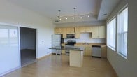This single-floor space showcases a thoughtfully designed open-concept layout that seamlessly blends the kitchen and living areas. The kitchen is a standout feature, equipped with a full suite of stainless steel appliances, including a refrigerator, electric range, microwave, and dishwasher. Light maple cabinetry lines the walls, providing ample storage, while dark granite countertops offer a striking visual contrast and generous workspace. A central kitchen island, also topped with granite, serves as a functional hub ideal for meal preparation or casual dining. Overhead, a combination of pendant lights and a track lighting system illuminates the space with layered, adjustable lighting. The flooring throughout is light hardwood, contributing to the bright and airy atmosphere of the interior. A tray ceiling detail above the kitchen area adds an architectural dimension and sense of depth. The open flow between the kitchen and the adjacent living area creates a spacious and versatile environment. The neutral color palette of the walls and cabinetry keeps the space feeling clean, modern, and welcoming throughout.