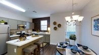 This two-story residence showcases a thoughtfully designed open-concept layout that seamlessly connects the kitchen and dining areas. The kitchen is a standout feature, equipped with gray-toned cabinetry, a contrasting dark wood accent wall, and white countertops that provide a clean, contemporary aesthetic. Stainless steel appliances, including an over-the-range microwave and a gas range, enhance the functional appeal. A peninsula with bar seating offers additional prep space and casual dining options, complemented by upholstered bar stools.

The adjacent dining area is anchored by a round table beneath an elegant multi-arm chandelier, creating an inviting atmosphere for meals and gatherings. Rich, dark hardwood-style flooring runs throughout both spaces, adding warmth and visual continuity. Multiple windows dressed with white blinds allow generous natural light to fill the area, brightening the neutral color palette of white walls and trim. Decorative wall plates and lush greenery add subtle personality to the space. The two-story configuration offers additional living areas beyond what is immediately visible, suggesting a spacious and versatile floor plan.