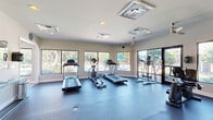This fitness center at 8100 U.S. 98 in Pensacola, Florida, offers a dedicated exercise room designed to support a comprehensive workout experience. The space is thoughtfully laid out with a durable rubber flooring surface that covers the entire workout area, providing both comfort and safety during exercise.

The room is generously outfitted with cardio equipment, including multiple treadmills and an elliptical machine, alongside a well-rounded selection of strength training stations and cable machines, as well as a stationary bike. Ceiling fans and overhead HVAC units ensure consistent airflow and climate control throughout the space.

Large windows line the perimeter walls, flooding the interior with natural light and offering views of the surrounding outdoor area, including what appears to be a pool area with mature trees. Glass-paneled doors provide additional access points and contribute to the bright, open atmosphere of the room.

A small entryway area serves as a transition into the fitness floor, featuring a wall-mounted TV, a glass-topped table, and vending machines for added convenience. Recessed lighting and ceiling-mounted fixtures supplement the natural light, keeping the space well-illuminated at all hours. The clean, neutral color palette of white walls and light ceilings reinforces a focused and energizing environment suited for fitness and wellness activities.