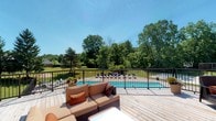 This single-story property showcases a well-appointed outdoor living area centered around a spacious elevated wooden deck. The deck is finished with light-toned hardwood planking and enclosed by a sleek black metal railing, providing both safety and unobstructed sightlines to the grounds below. Comfortable wicker sectional seating with tan cushions and accent pillows anchors the deck space, creating an inviting lounge area perfect for entertaining or unwinding.

From the deck, residents enjoy a direct view of a generously sized rectangular swimming pool situated on the lower level of the property. The pool area is lined with lounge chairs, suggesting ample space for sunbathing and recreation. A well-maintained lawn surrounds the pool, extending outward to a dense border of mature deciduous trees that provide natural privacy and a lush green backdrop throughout warmer months.

The overall ambiance of the outdoor space is one of openness and tranquility, enhanced by abundant natural light and the sense of seclusion offered by the surrounding landscape. The thoughtful layering of the elevated deck above the pool area creates a dynamic outdoor environment well-suited for both relaxation and social gatherings.