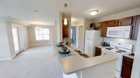 This single-floor residence offers a thoughtfully designed open-concept layout that seamlessly connects the kitchen and living areas, creating a spacious and functional environment. The kitchen is a standout feature, anchored by a generous peninsula with a built-in double sink and a light-toned countertop that comfortably accommodates bar seating. Rich, warm-toned cabinetry provides ample storage, while white appliances, including a full-size refrigerator and range, lend a clean, classic look. A stylish pendant light hangs above the peninsula, adding a decorative focal point to the space. The kitchen flows naturally into the open living area, which is finished with plush wall-to-wall carpeting that adds warmth and comfort underfoot. Natural light filters in through a large window adorned with classic white trim, brightening the living space and enhancing its airy feel. An elegant chandelier overhead adds a touch of sophistication to the dining and living zone. Neutral wall tones throughout create a versatile backdrop, making the space easy to personalize. Overall, this residence combines practical design with comfortable, welcoming finishes suited to everyday living.