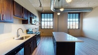 This single-floor urban loft blends industrial character with contemporary finishes to create a striking and functional living space. The open-concept layout is anchored by soaring ceilings with exposed ductwork and corrugated metal paneling, lending an authentic warehouse aesthetic throughout. Large, multi-pane factory-style windows flood the interior with abundant natural light while framing panoramic city views, reinforcing the loft's urban character.

The kitchen is a standout feature, outfitted with rich dark cabinetry, a full suite of stainless steel appliances including a refrigerator, range, microwave, and dishwasher, and a spacious island topped with a crisp white countertop. Pendant lighting with an industrial flair hangs above, complementing the overall design. Floating wood shelves add both style and practicality to the kitchen wall.

The living area is open and expansive, defined by light wood-tone flooring that runs throughout the main space. A distinctive decorative column adds architectural interest and speaks to the building's historic bones. A glass-paneled partition provides a subtle room division while maintaining visual openness.

The bathroom is clean and modern, featuring large-format tile flooring, a glass-enclosed walk-in shower with tiled surround, and a vanity with ample storage. A dedicated utility area houses a stacked in-unit washer and dryer, adding everyday convenience to this well-appointed urban retreat.