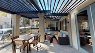 The outdoor patio at Arroyo Villa Apartments is a thoughtfully designed communal space that blends architectural structure with comfortable, resort-style amenities. The area is anchored by a bold, dark-stained pergola overhead, its evenly spaced horizontal beams allowing filtered natural light and open sky views while defining the space beneath. Stucco-finished columns with decorative detailing support the pergola structure and complement the building's exterior aesthetic.

The patio floor is laid with interlocking stone pavers in a neutral tone, providing a durable and visually appealing surface throughout. A large sectional sofa with patterned blue and neutral cushions, accented with orange throw pillows, creates an inviting lounge area arranged around low accent tables. Nearby, a wooden dining table with matching chairs offers a dedicated outdoor eating space.

Large sliding glass windows along the building's facade connect the patio visually to the interior, while wall-mounted lantern-style light fixtures provide ambient illumination for evening use. Teal-framed glass entry doors provide access between the indoor and outdoor areas. The overall ambiance is polished and welcoming, suitable for relaxed social gatherings.