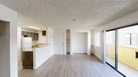 This single-floor unit offers a practical, open-concept layout highlighted by light wood-look flooring that flows throughout the living areas. The living room is spacious and bright, featuring a sliding glass door that opens to a private balcony, allowing natural light to fill the space. A wall-mounted HVAC unit provides climate control, and vertical blinds offer privacy. Built-in shelving units on either side of the main doorway provide convenient storage and display options.

The kitchen is positioned at one end of the open layout, featuring dark brown cabinetry, a double stainless steel sink set into a light-toned countertop, a white refrigerator, and a freestanding electric range. A peninsula counter separates the kitchen from the living area, making it ideal for casual dining or meal prep. A textured popcorn ceiling is consistent throughout the unit.

The bedroom includes a sliding-door closet offering ample hanging and storage space. The bathroom is straightforward and functional, with a single-sink dark wood vanity with a countertop, a tub/shower combination with a curtain rod, and wood-look flooring that mirrors the rest of the unit. The overall aesthetic is clean, neutral, and move-in ready.