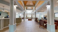 This single-floor residence offers a thoughtfully arranged layout that balances open communal spaces with private living areas. The living room serves as the centerpiece of the unit, featuring neutral-toned carpet, warm beige walls, and a textured ceiling. Multiple windows fitted with white horizontal blinds allow natural light to filter throughout the space, creating a bright and airy atmosphere. A door provides direct exterior access from the living area. A half-wall partition with a dark countertop separates the living room from the kitchen, creating a semi-open concept feel while maintaining a defined cooking zone.

The kitchen is equipped with rich medium-toned wood cabinetry, dark countertops, and black appliances, including an electric range and an over-the-range microwave. Light hardwood-style flooring grounds the kitchen, contrasting with the carpeted areas of the unit. Storage is well-represented through the generous upper and lower cabinet configuration.

The bedroom continues the neutral color palette with beige walls and plush carpet underfoot. A wide bifold closet provides ample storage, while a window brings in natural light. The overall design of the space is clean, functional, and move-in ready, offering a comfortable and practical living environment throughout.