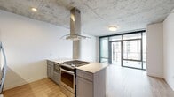 This thoughtfully designed one-bedroom, one-bathroom unit showcases a contemporary industrial aesthetic throughout its efficiently organized single-floor layout. The open-plan kitchen and living area serves as the heart of the space, anchored by a striking kitchen island equipped with a stainless steel gas cooktop, a built-in oven, and a dramatic stainless steel island range hood that extends to the exposed concrete ceiling above. Gray shaker-style cabinetry, quartz countertops, a subway tile backsplash, and a full suite of stainless steel appliances complete the well-appointed kitchen.

The living area flows seamlessly toward floor-to-ceiling glass doors that open to a private balcony, flooding the space with natural light and offering expansive urban views. Light wood-tone flooring runs continuously throughout, complementing the raw texture of the exposed concrete ceiling—a defining architectural feature of the unit.

The generously sized primary bedroom benefits from its own set of floor-to-ceiling windows with sweeping city views and includes access to a dedicated closet. An exposed concrete column adds to the industrial character of the room. The bathroom is finished with large-format tile flooring, gray shaker vanity cabinetry, a white quartz countertop, and a frameless glass walk-in shower with subway tile surround.