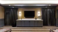 This single-floor space exudes a polished, contemporary aesthetic designed to impress guests and facilitate a variety of gatherings. The interior is anchored by a striking feature wall finished in warm gold tones, set against deep charcoal and dark drapery that frames the space with a sense of refined drama. A large flat-screen display is wall-mounted at the center of this feature wall, flanked by stylish sconce lighting that casts a warm, inviting glow. Below the display, a generous built-in credenza with sleek grey cabinetry and a crisp white countertop provides ample storage and display surface, thoughtfully accessorized with decorative vases and accent pieces. The flooring features a patterned carpet with horizontal striping in neutral and gold tones, adding texture and visual interest underfoot while complementing the overall color palette. Dark ceiling elements and controlled ambient lighting contribute to an intimate yet upscale atmosphere throughout. The overall design is cohesive and purposeful, balancing functionality with an elevated aesthetic that makes this space well-suited for presentations, social events, or private gatherings.