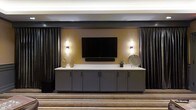 This single-floor space exudes a polished, contemporary aesthetic designed to impress guests and facilitate a variety of gatherings. The interior is anchored by a striking feature wall finished in warm gold tones, set against deep charcoal and dark drapery that frames the space with a sense of refined drama. A large flat-screen display is wall-mounted at the center of this feature wall, flanked by stylish sconce lighting that casts a warm, inviting glow. Below the display, a generous built-in credenza with sleek grey cabinetry and a crisp white countertop provides ample storage and display surface, thoughtfully accessorized with decorative vases and accent pieces. The flooring features a patterned carpet with horizontal striping in neutral and gold tones, adding texture and visual interest underfoot while complementing the overall color palette. Dark ceiling elements and controlled ambient lighting contribute to an intimate yet upscale atmosphere throughout. The overall design is cohesive and purposeful, balancing functionality with an elevated aesthetic that makes this space well-suited for presentations, social events, or private gatherings.
