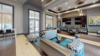 Located at 3115 Grand Ave in Pinellas Park, Florida, this contemporary single-story clubhouse offers a thoughtfully designed gathering space with a polished, upscale aesthetic. Upon entry, visitors are greeted by a bright foyer featuring large floor-to-ceiling windows fitted with warm roller shades, bold abstract artwork, and sleek leather seating — setting an immediately welcoming tone. The flooring transitions elegantly between light tile and rich wood-look planks throughout the space.

The main lounge area is anchored by a coffered ceiling with recessed and pendant lighting, creating a layered ambiance. Comfortable modern sofas and colorful patterned ottomans are arranged around a central coffee table, while a full bar and dining area with counter seating and a striking mosaic tile backsplash occupies the far end of the room. Large windows and glass double doors flood the interior with natural light and provide views of the palm-lined exterior.

A dedicated recreation area features a billiards table, dual wall-mounted flat-screen televisions on a built-in display shelf, and dark wood cabinetry, offering a more casual entertainment setting. Throughout, the design palette of cool grays, teal accents, and dark wood finishes gives the space a cohesive, contemporary character well-suited for community gatherings and social events.