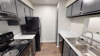Located at 7674 Courtney Oaks Dr in Fort Worth, TX, this single-floor unit offers a thoughtfully laid-out living space with a clean, neutral palette throughout. The galley-style kitchen is well-appointed with gray cabinetry, light speckled countertops, a stainless steel sink, and black appliances including a refrigerator, electric range, and dishwasher. Wood-look flooring runs through the kitchen and entry areas, adding a warm, cohesive aesthetic. The open living area transitions from wood-look flooring to plush carpeting, creating distinct zones within the space. A ceiling fan provides comfort, and vertical blinds along the exterior wall allow for natural light control. The bedroom is generously sized, featuring soft carpeting, a ceiling fan with light fixture, and a window fitted with horizontal blinds that invite natural light. A walk-in closet with built-in shelving provides ample storage. The full bathroom is finished with wood-look flooring, a dark vanity with a speckled countertop, a tub-shower combination, and brushed nickel hardware and towel bars. Textured ceilings are present throughout the unit, contributing to the overall character of the space.