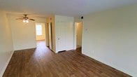 This single-floor residence offers a clean, functional layout designed for comfortable everyday living. The main living area features warm, rich hardwood-style flooring that extends throughout the space, complementing the soft, neutral cream-toned walls that reflect natural light and create an open, airy atmosphere. A central dividing wall with a white-paneled closet door provides a natural separation between spaces while maintaining an open feel.

Multiple doorways lead to additional rooms, offering a well-connected floor plan. Natural light filters in from a window visible in an adjoining room, brightening the interior and enhancing the sense of spaciousness. HVAC vents are positioned along the ceiling and walls, ensuring climate comfort throughout. White trim and door frames provide clean contrast against the warm flooring and neutral walls.

The layout includes storage in the form of a built-in closet, contributing to the practical design of the space. The overall ambiance is understated and move-in ready, presenting a blank canvas well-suited for a variety of furnishing styles. The consistent flooring material and cohesive color scheme throughout give the home a unified, polished appearance.