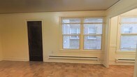This single-floor space offers a straightforward and functional layout with a number of notable architectural details. The main living area features classic parquet wood flooring arranged in a distinctive pattern, adding warmth and character to the room. A large double-pane window fitted with slatted blinds allows natural light to filter into the space, with views looking out toward an adjacent brick building and a walkway below. A baseboard heating unit runs along the wall beneath the window, providing efficient and even heat distribution throughout the room. The walls are finished in a neutral tone with decorative crown molding detail framing the upper perimeter, contributing a traditional aesthetic to the interior. A dark-stained wooden door is visible to one side, offering clean contrast against the lighter walls. The overall ambiance is understated and practical, with classic design elements that provide a solid foundation for personalization.