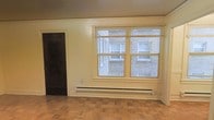 This single-floor space offers a straightforward and functional layout with a number of notable architectural details. The main living area features classic parquet wood flooring arranged in a distinctive pattern, adding warmth and character to the room. A large double-pane window fitted with slatted blinds allows natural light to filter into the space, with views looking out toward an adjacent brick building and a walkway below. A baseboard heating unit runs along the wall beneath the window, providing efficient and even heat distribution throughout the room. The walls are finished in a neutral tone with decorative crown molding detail framing the upper perimeter, contributing a traditional aesthetic to the interior. A dark-stained wooden door is visible to one side, offering clean contrast against the lighter walls. The overall ambiance is understated and practical, with classic design elements that provide a solid foundation for personalization.