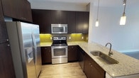 This single-floor residence showcases a thoughtfully designed kitchen that serves as a central highlight of the space. The kitchen features rich, dark espresso-toned cabinetry that extends floor to ceiling, maximizing storage while lending a sophisticated, contemporary aesthetic. Light-veined granite countertops provide a striking contrast against the dark cabinets, offering generous prep and workspace throughout.

The kitchen is equipped with a full suite of stainless steel appliances, including a French door refrigerator, an electric range with oven, an over-the-range microwave, and a dishwasher — providing everything needed for comfortable, efficient cooking. A yellow-green tile backsplash adds a bold pop of color and visual interest between the countertops and upper cabinetry. Under-cabinet lighting subtly illuminates the workspace, enhancing both functionality and ambiance.

Light wood-tone flooring runs throughout, complementing the overall modern palette and contributing to the bright, open feel of the space. A pendant light fixture is visible near the sink area, adding a refined decorative touch. The layout is designed for practical flow, with ample counter space along multiple walls and a deep stainless steel sink with a modern faucet completing the well-curated design.