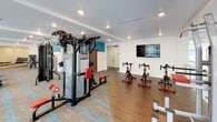 This contemporary fitness center is thoughtfully designed to support a comprehensive workout experience. The space is bright and open, benefiting from large windows along the perimeter that flood the interior with natural light and offer outdoor views, creating an energizing atmosphere for exercise.

The flooring combines patterned carpet tiles in earthy tones with teal accents alongside hardwood-style sections, adding visual interest and defining distinct workout zones throughout the space. Recessed lighting in the white ceiling provides consistent illumination throughout.

A generous row of cardio equipment lines one side of the space, featuring multiple treadmills and elliptical machines. On the opposite side, a multi-station cable machine serves as a centerpiece for strength training, accompanied by additional selectorized weight machines and benches for targeted muscle work. A dedicated indoor cycling area houses several stationary spin bikes positioned near a wall-mounted flat-screen television. 

Toward the rear, a mirrored free weights section provides a full rack of dumbbells and a workout bench, ideal for functional training. Vibrant teal accent walls punctuate the otherwise clean white interior, adding a bold, modern aesthetic. A private room with a wood-panel door is also accessible from the main floor.