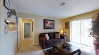 This single-floor residential space offers a welcoming and functional layout designed for comfortable everyday living. The living room serves as the heart of the home, featuring wall-to-wall carpeting that adds warmth underfoot and a soft, neutral color palette across the walls that creates an inviting ambiance. A large sliding glass door with vertical blinds allows an abundance of natural light to fill the room, brightening the space throughout the day. The living area comfortably accommodates a plush sofa seating arrangement centered around a glass-top coffee table, making it ideal for relaxing or entertaining. Decorative wall art adds a touch of personality to the space. A textured ceiling adds architectural character overhead, complemented by a central HVAC vent for climate control. The living room opens directly into a well-lit hallway, providing seamless access to the rest of the unit. The hallway features the same light carpeting and connects to additional rooms, maintaining visual continuity throughout. Overall, the space presents a cozy, approachable atmosphere with practical design elements suited for day-to-day residential living.