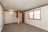 This single-floor space offers a practical and straightforward layout connecting several distinct living areas. The living room is generously sized, featuring neutral wall-to-wall carpeting, white walls accented by a decorative border near the ceiling, and a wood-trimmed front door alongside a large window that allows natural light to filter into the space. An adjacent carpeted room mirrors a similar aesthetic, also featuring a window, ceiling vent, and a wall-mounted light fixture, making it suitable as a bedroom or secondary living space.

The kitchen is efficiently arranged in an L-shaped configuration, finished with light ceramic tile flooring. It features warm-toned wood cabinetry above and below the counters, laminate countertops with a stainless steel sink, a white electric coil-burner range with an overhead exhaust hood, and a white refrigerator. Ample cabinet storage lines the walls, providing functional workspace throughout.

The bathroom carries a distinctly retro character, highlighted by vintage teal-colored fixtures including the bathtub and toilet, complemented by matching teal ceramic wall tiles. A window above the tub provides natural ventilation, and a chrome towel bar and shower curtain rod complete the space. Overall, the unit presents a classic, functional interior with consistent design elements throughout.
