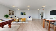 Located at 400 N Main St in Glen Ellyn, Illinois, this single-floor community amenity space is designed with a clean, contemporary aesthetic that prioritizes comfort and social interaction. Light wood-look flooring runs throughout, complementing the crisp white walls and modern flush-mount lighting that keep the interiors bright and welcoming.

The expansive lounge area serves as the heart of the space, furnished with generous white sofas, warm wood-framed accent chairs, and layered area rugs that create an inviting atmosphere for relaxation or casual gatherings. A large-screen television is wall-mounted for entertainment, and curated artwork adds personality to the open layout.

Adjacent to the lounge, a dedicated billiards room offers a full-sized pool table set beneath large, black-framed floor-to-ceiling windows that frame views of the surrounding Glen Ellyn streetscape. Glass-paneled double doors provide direct access to an outdoor area, flooding the room with natural light.

The kitchen and dining area is outfitted with white cabinetry, subway tile backsplash, stainless steel appliances, and a generous high-top dining table with seating for multiple guests. A large window brings in ample daylight and offers outdoor views, making this a functional and pleasant space for communal dining or casual meetings. Together, these areas form a well-appointed, multipurpose community retreat.