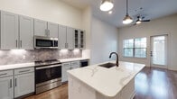 This single-story space showcases a thoughtfully designed open layout with high ceilings that enhance the sense of volume throughout. The kitchen serves as a central highlight, featuring a large waterfall-edge quartz island with an undermount sink and matte black faucet. Gray shaker-style cabinetry extends to the upper walls, complemented by a herringbone-patterned tile backsplash with under-cabinet lighting. Stainless steel appliances, including a French door refrigerator, range, and built-in microwave, provide a polished, modern aesthetic. Pendant lighting and a ceiling fan complete the kitchen and adjoining living area.

Hardwood-style flooring flows through the main living and kitchen areas, while plush carpet adds comfort to the bedroom, which benefits from generous natural light through large, blinds-fitted windows. A glass-paned door in the living area provides outdoor access and additional light.

The bathroom is well-appointed with a dual-sink vanity featuring gray cabinetry, white quartz countertops, matte black fixtures, a large framed mirror, wood-look flooring, and a combination tub and shower with dark tile surround. The overall aesthetic is clean, contemporary, and cohesive.
