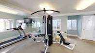 Located at 3105 South 47th Street in Tacoma, Washington, this fitness center offers a well-equipped, single-floor exercise facility designed to support a comprehensive workout experience. The space is finished with soft blue-gray walls, white trim, and neutral carpet throughout, creating a clean and inviting atmosphere. Large windows on multiple walls allow generous natural light to fill the room, enhancing the open, airy feel of the space.

The fitness center is outfitted with a diverse selection of equipment to accommodate both cardio and strength training needs. Cardio offerings include a True treadmill, an elliptical trainer, and a stair climber, all positioned to take advantage of the natural light from the surrounding windows. A multi-station cable strength machine serves as the centerpiece for resistance training, complemented by a weight bench and free weights for additional versatility.

A full-length wall mirror runs along one side of the room, visually expanding the space and providing users with proper form feedback during workouts. A wall-mounted television is also present, adding an entertainment option during exercise sessions. A water fountain is conveniently located within the space, and a fire safety station is accessible near the entrance. Flush-mounted ceiling light panels provide consistent, even illumination throughout. The space is accessible via multiple entry points, making it convenient and functional for regular use.