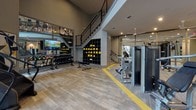 This contemporary fitness center is thoughtfully designed to support a comprehensive workout experience. The space is anchored by soaring double-height ceilings in the main training area, with large windows that flood the floor with natural light and provide views to the outdoors. Exposed ductwork and industrial-style ceiling details add a modern edge to the overall aesthetic.

The main floor is organized into distinct workout zones, clearly delineated by bold yellow floor striping on durable wood-look flooring. The strength training area is well-stocked with cable machines, a multi-function rack, weight benches, barbells, and a stair climber. A dedicated free weights section includes a full dumbbell rack, kettlebells in various sizes, and a flat bench, accompanied by a large wall-mounted display screen for entertainment or instructional use.

Floor-to-ceiling mirrors line portions of the walls, expanding the sense of space and supporting form-focused training. Cubbies for personal items and accessory storage are built into the wall near the base of the staircase leading to the mezzanine level above. The lower level also features additional cable and resistance machines, ceiling fans for comfort, and modern artwork that adds a polished, welcoming atmosphere throughout.
