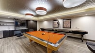 This well-appointed game room delivers a refined entertainment experience through a thoughtfully designed single-floor layout. At the center of the space stands a full-size pool table with a bold orange felt surface and a solid wood frame, serving as the room's focal point. Overhead, a decorative geometric-patterned ceiling tile adds a distinctive architectural touch, complemented by a coffered tray ceiling with warm ambient uplighting and large drum pendant light fixtures that create an inviting atmosphere.

Along one wall, a full-length shuffleboard table provides additional gaming options, accompanied by framed billiards-themed artwork that reinforces the room's recreational character. A wet bar area features dark wood cabinetry, a mosaic tile backsplash, and a mounted flat-screen television, making it ideal for refreshments and casual viewing. Modern black leather lounge chairs and small side tables are arranged throughout, offering comfortable seating for guests.

Light wood-look flooring runs throughout the space, tying together the contemporary design elements. Natural light filters in through horizontal windows along one wall, while a warm-toned glass-paneled entry door adds an elegant touch to the entrance area. The overall ambiance is sleek, modern, and well-suited for social gatherings and recreational use.