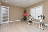 Located at 700 SR-96 E in Bonaire, Georgia, this single-floor fitness studio is thoughtfully designed to support a full range of exercise activities. The space features light hardwood flooring throughout, creating a clean and energizing atmosphere complemented by warm, neutral-toned walls and recessed lighting that ensures bright, even illumination across the room.

The main workout area is generously proportioned and equipped with multiple stationary cycling bikes, a tiered dumbbell rack with color-coded weights, stability balls, and a rolled exercise mat — offering versatility for both cardio and strength training. A large wall-mounted flat-screen display is prominently positioned, making it well-suited for streaming virtual fitness classes or instructional content.

A framed window allows natural light to filter into the space, enhancing the open and welcoming feel of the room. On one side, a full-height glass-paneled door with a modern multi-lite design provides both style and transparency, connecting the studio to an adjoining corridor with rich hardwood flooring. Recessed ceiling speakers suggest built-in audio capability, further supporting an immersive workout environment.

The studio's clean lines, functional layout, and quality finishes make it a practical and polished space for fitness instruction, personal training, or community exercise programming.