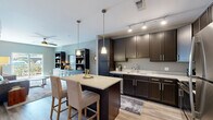 This single-floor residence offers a well-planned open-concept layout that seamlessly connects the kitchen, dining, and living areas. The kitchen is a standout feature, outfitted with rich dark-espresso cabinetry, light granite countertops, stainless steel appliances, and a central island with bar seating illuminated by elegant pendant lighting. Track lighting overhead adds a polished, modern touch, while light wood-style flooring ties the space together with warmth and continuity.

The living room is bright and inviting, benefiting from natural light through a sliding glass door that opens to an outdoor area. A built-in shelving and cabinetry unit in deep charcoal anchors the room, providing both display and storage space. A ceiling fan adds comfort to the relaxed, contemporary atmosphere.

The bedroom is a calm and comfortable retreat, featuring plush carpet, a ceiling fan, and a window dressed with sheer curtains that allow natural light to filter through. A bold accent wall adds a touch of personality to the space.

The bathroom is clean and functional, featuring subway tile surrounding the tub-shower combination, a dark-framed vanity mirror, granite countertops, and durable wood-look flooring. Adjacent wire-rack shelving provides ample organizational storage, complementing the overall practical elegance of this well-appointed residence.