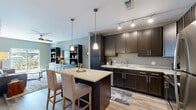 This single-floor residence offers a well-planned open-concept layout that seamlessly connects the kitchen, dining, and living areas. The kitchen is a standout feature, outfitted with rich dark-espresso cabinetry, light granite countertops, stainless steel appliances, and a central island with bar seating illuminated by elegant pendant lighting. Track lighting overhead adds a polished, modern touch, while light wood-style flooring ties the space together with warmth and continuity.

The living room is bright and inviting, benefiting from natural light through a sliding glass door that opens to an outdoor area. A built-in shelving and cabinetry unit in deep charcoal anchors the room, providing both display and storage space. A ceiling fan adds comfort to the relaxed, contemporary atmosphere.

The bedroom is a calm and comfortable retreat, featuring plush carpet, a ceiling fan, and a window dressed with sheer curtains that allow natural light to filter through. A bold accent wall adds a touch of personality to the space.

The bathroom is clean and functional, featuring subway tile surrounding the tub-shower combination, a dark-framed vanity mirror, granite countertops, and durable wood-look flooring. Adjacent wire-rack shelving provides ample organizational storage, complementing the overall practical elegance of this well-appointed residence.