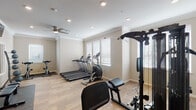 Located at 597–613 W Henri de Tonti Blvd in Tontitown, Arkansas, this single-floor fitness center is thoughtfully designed to support a comprehensive workout experience. The space features light wood-style flooring throughout, neutral warm-toned walls, and crown molding that lends a polished, refined aesthetic uncommon in typical gym environments.

The cardio zone is well-appointed with multiple treadmills, an elliptical trainer, and a stationary cycle, all positioned near large windows fitted with horizontal blinds that allow ample natural light to fill the room. Ceiling fans with recessed lighting ensure consistent airflow and bright illumination throughout the space.

The strength training area includes a multi-station cable machine, a dedicated dumbbell rack with a full range of free weights, a weight bench with mat, and additional resistance equipment. Full-length wall mirrors are framed in white and positioned to allow users to monitor their form during exercises. Multiple flat-screen TVs are wall-mounted above the mirrors, providing entertainment during workouts.

A built-in dark wood cabinetry unit with a stone countertop is integrated into one wall, offering practical storage and surface space. The overall layout is open and well-organized, creating an inviting, functional fitness environment suited for a variety of training routines.