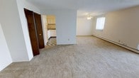 This single-floor residential unit offers a thoughtfully arranged layout that maximizes functionality and comfort. The open living area is a standout feature, finished with wall-to-wall carpeting that adds warmth underfoot. A ceiling fan overhead and a baseboard heater along the wall ensure year-round comfort, while a bright window allows natural light to fill the space. The living area flows seamlessly into the kitchen, which is distinguished by its classic black-and-white checkered tile flooring — a timeless design element that gives the space character. The kitchen is outfitted with white cabinetry, a refrigerator, and a sink set beneath a tiled backsplash, providing a clean and efficient cooking environment. The floor plan, as seen in the dollhouse view, reveals a well-organized series of rooms connected by interior doors, including what appears to be a bathroom and additional private spaces. Closet access is conveniently integrated throughout. The neutral color palette of white and soft grey across the walls creates a bright, airy ambiance that makes the unit feel welcoming and easy to personalize. Overall, the space combines classic design touches with practical everyday living features.
